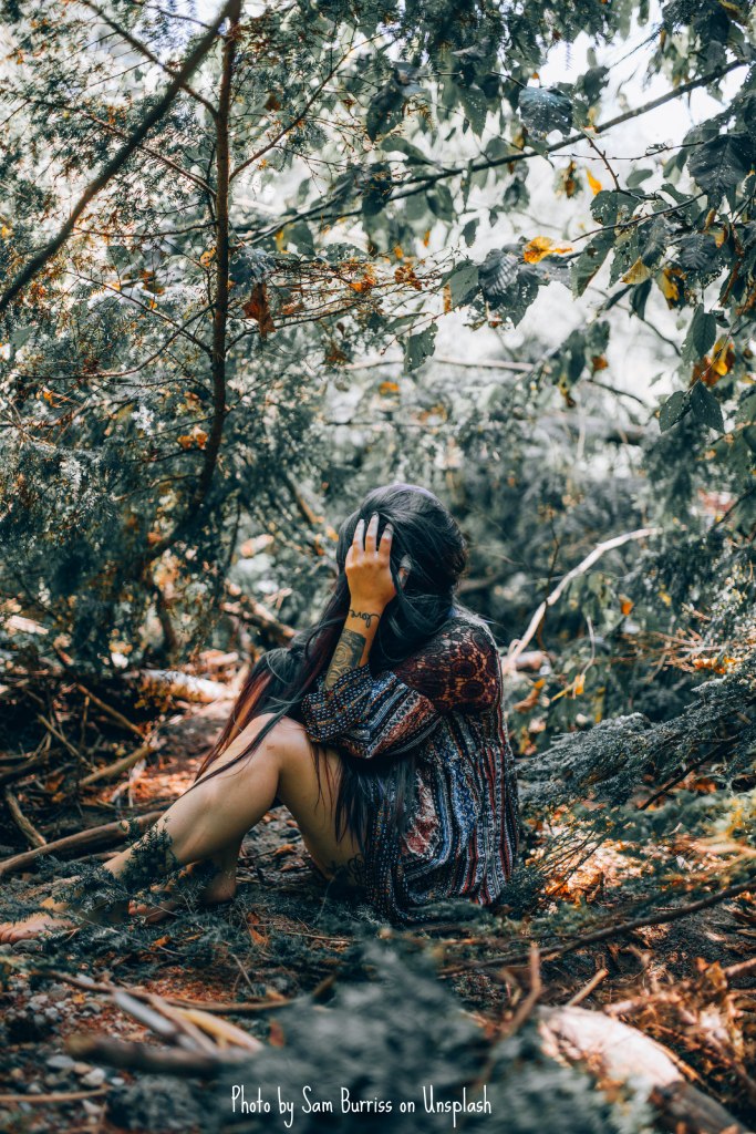 A person with long dark hair wearing a multi-colored long-sleeve dress is sitting on the ground in a forest. Branches from the nearby trees seem to be extending toward this person, who has their face buried in one hand, and a gap in the greenery behind the individual shows that there is much more forest to be explored. Judging by the various yellow and orange leaves in the trees and scattered on the forest floor, the photo seems to have been taken in the fall.