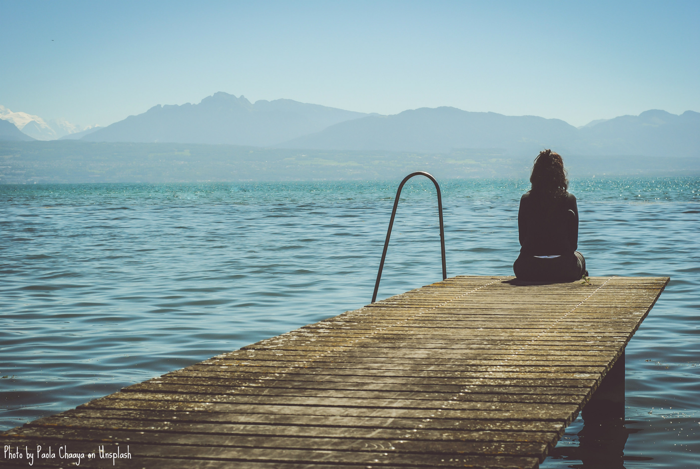 An individual dressed in dark clothing sits at the furthest end of a worn wooden dock. Grassy hills and mountain-like terrain are seen off in the distance. Their back is turned to us as they stare out across the dark water.