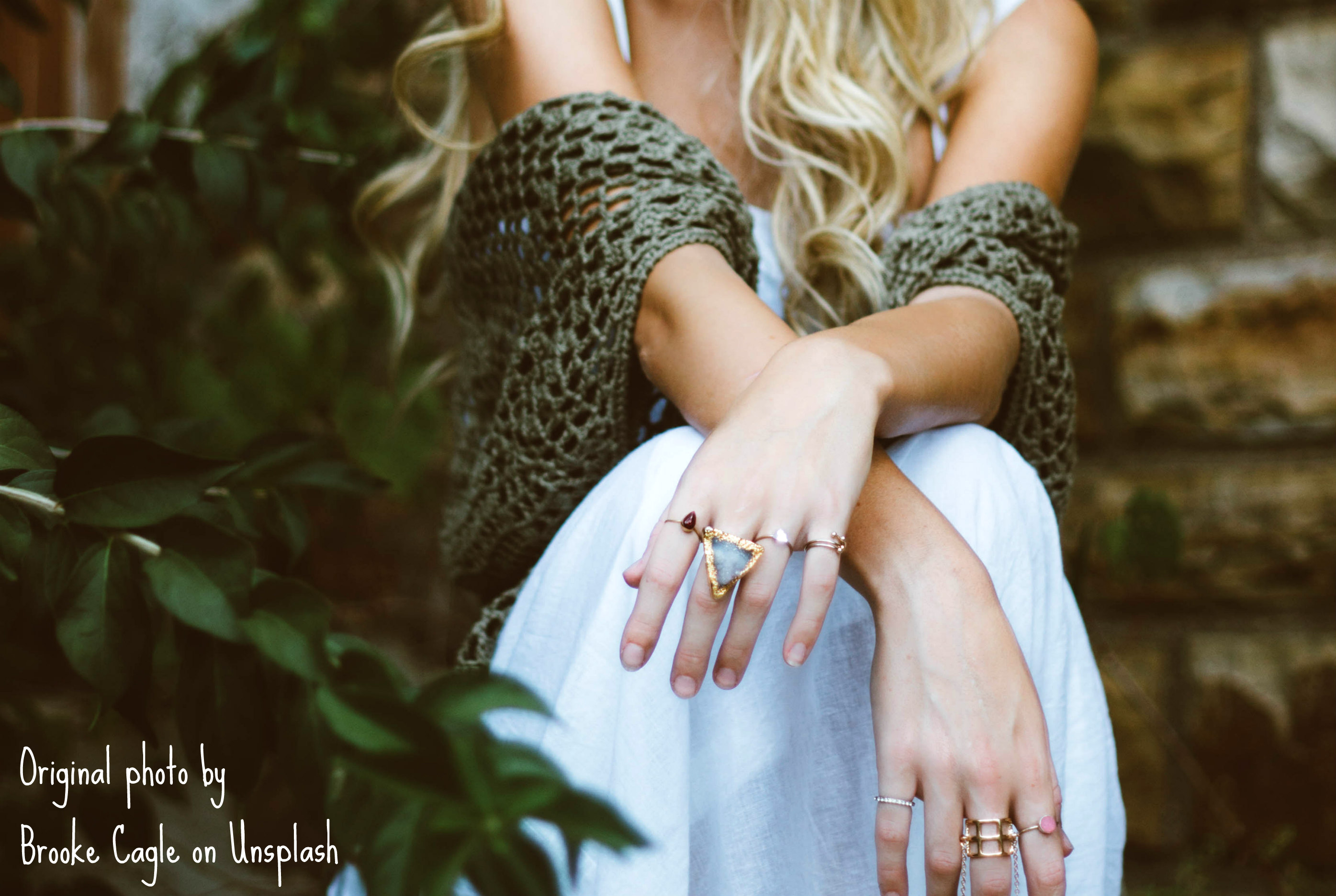 Person with long curly blonde hair in a white dress is seated in an area surrounded by green leaves, an olive shawl draped over their forearms as they lean their crossed wrists on their knees.