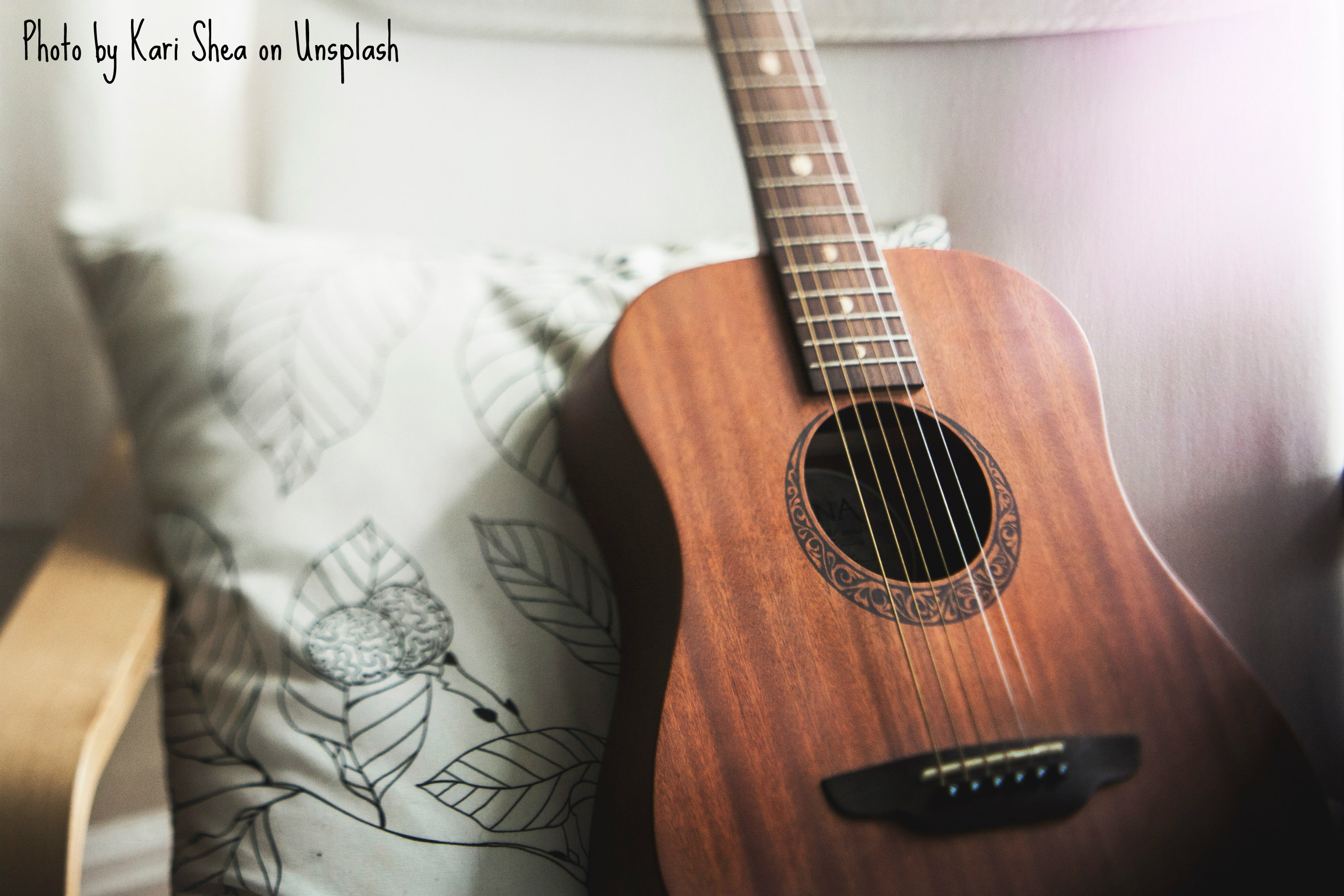 A six-stringed accoustic guitar is propped up against a white throw pillow that has leaves embroidered all over in black. A black crescent cups the sound hole, its fill pattern appearing to have a Celtic design. The crescent complements the mahogany body.