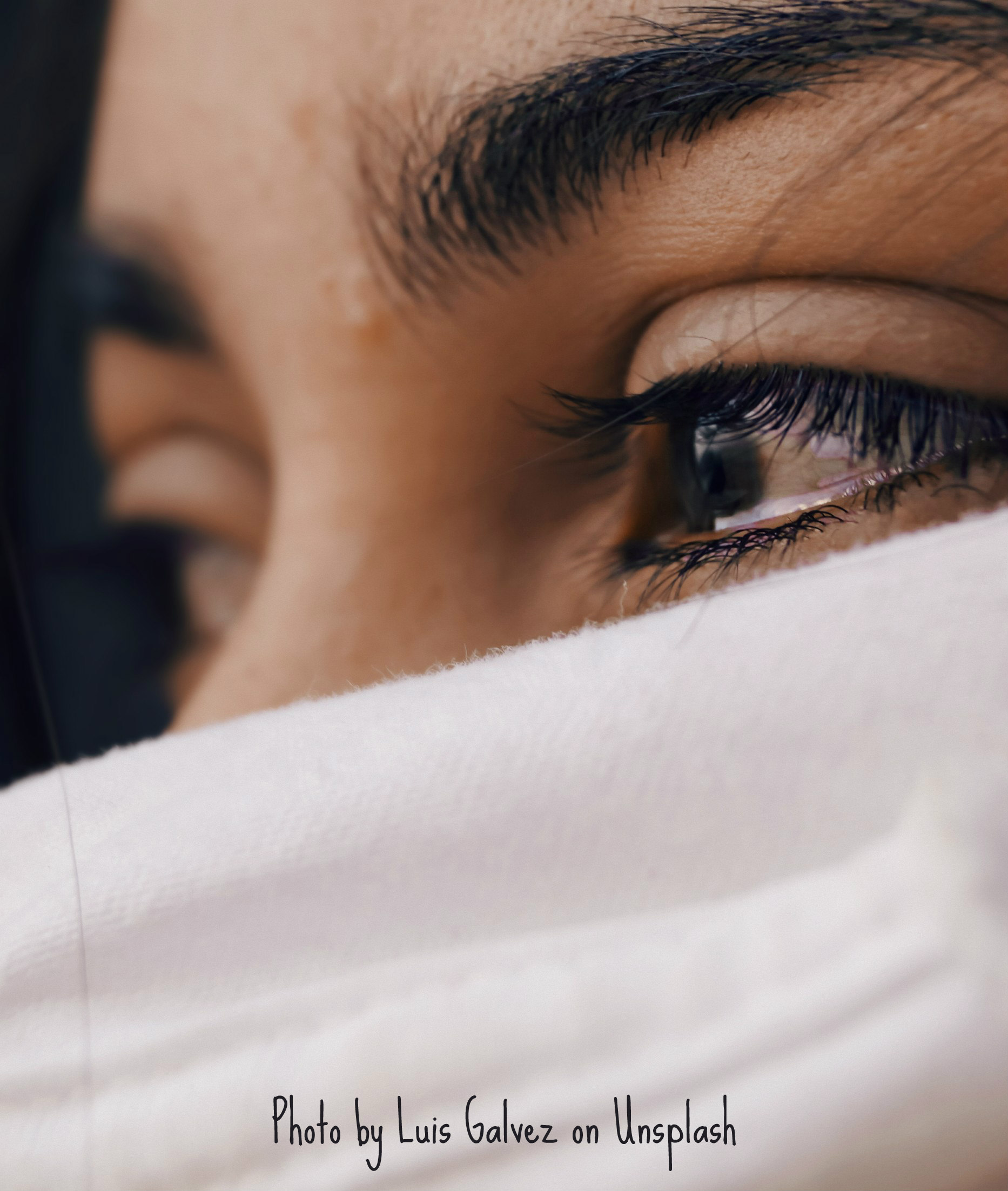 A close up of a person's face as they peer over what appears to be white fabric, presumably a blanket or article of clothing. Only their eyes and the upper bridge of their nose are visible from this left-side view. Both eyes appear discolored, possibly bloodshot, and the nearest one glistens with excess moisture, granting the notion that the model is distressed.