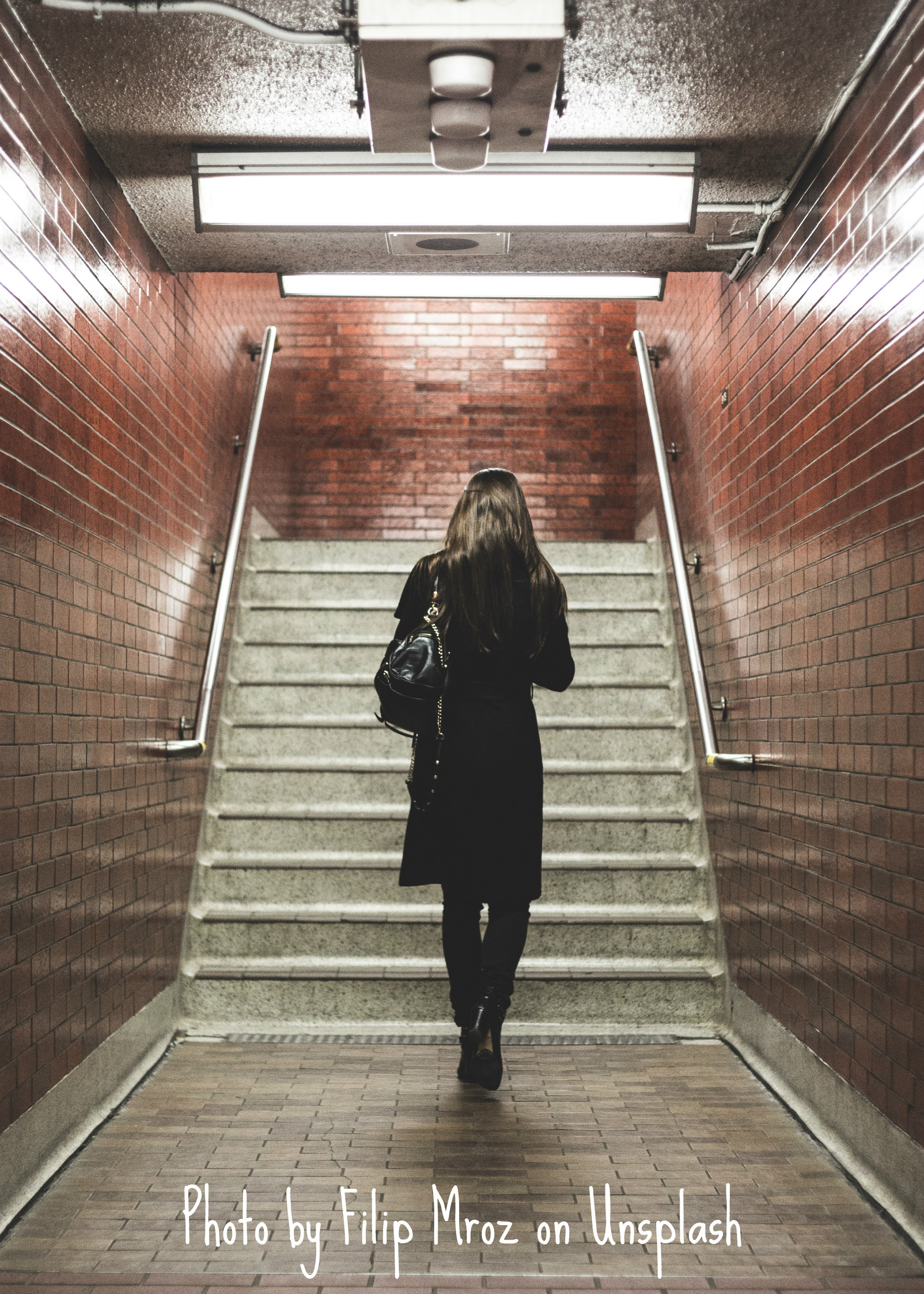 A lone individual with long, dark hair wearing a black trench coat, tights, and boots has their back turned and head lowered as they walk away. They are in a building or underground tunnel; the walls are lined with red brick, the ground is covered in square fray stone tiles, and long light fixtures hover overhead. Before the person is a set of stairs, metal rails on either side, with the next landing in sight and presumably veering off to the right. The model is about to ascend the steps.