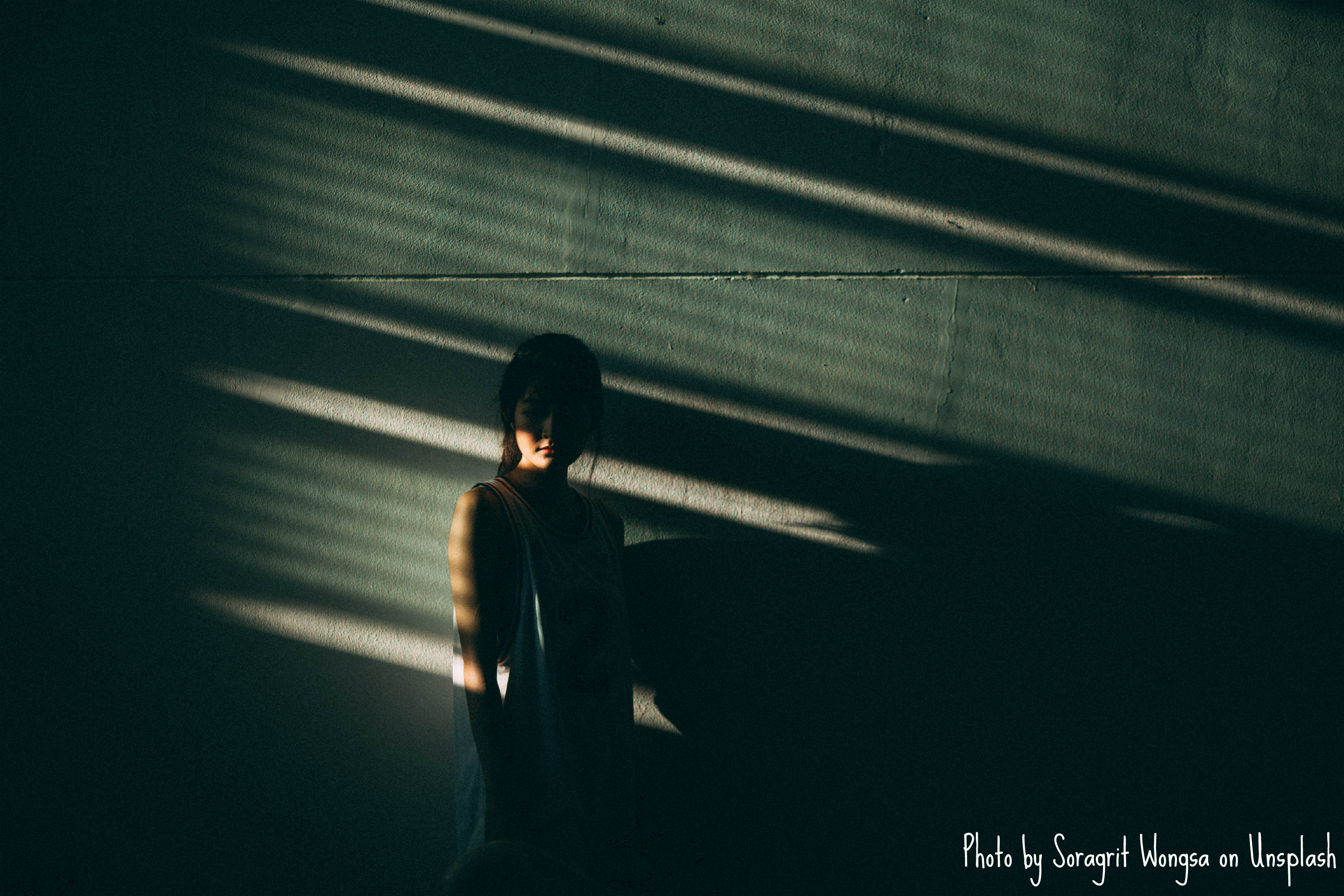 Petite person standing in a dark room, the cement walls allowing the presumption that the room is underground. Light filters through vertical gaps, leaving diagonal shadows on the individual and the wall. The top half of their face is obscured in darkness.