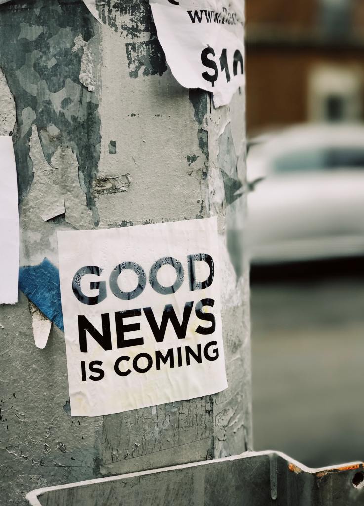 Wooden pole on the side of a road that has been painted white and has signs taped on it. The paint is peeling, revealing bluish-gray beneath, and one of the visible signs is ripped along the bottom. The main sign in focus is white with black font reading, "Good news is coming" in all capital letters. Out of focus in the background is a vehicle parked before a building. The building is red, presumably brick, and the car looks to be either white or silver. All the signs on the pole are wrinkled, with the main one showing signs of water damage.